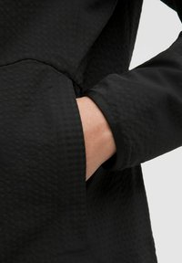 Černá bunda s texturovanou tkaninou a diagonálním vzorem. Detailní záběr ukazuje ruku v boční kapse, což zdůrazňuje design rukávu.