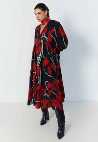 Zwarte jurk met rode bloemenprint en witte accenten, met een diepe V-hals en gelaagd ontwerp. Gecombineerd met zwarte knee-high laarzen.