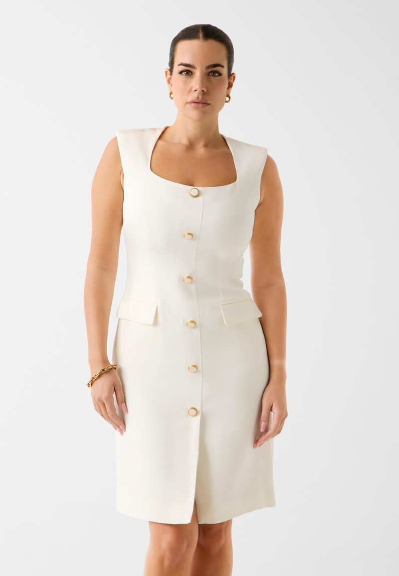 Femme portant une robe blanche sans manches avec des boutons dorés sur le devant, un décolleté carré et de petites poches à rabat, posant devant un fond uni.