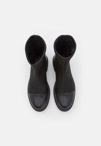 Un par de botas negras de tobillo con punteras de cuero suave y partes superiores de tela, vistas desde arriba sobre un fondo blanco.