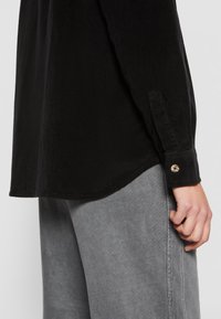Gros plan d'une personne portant une chemise noire à manches longues avec des poignets boutonnés et un pantalon gris, main reposant sur le côté.