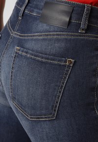 Mörka denimjeans har en slät yta, kontrasterande gyllene sömmar och en svart läderlapp med texten "CAMBIO JEANS" på midjebandet.