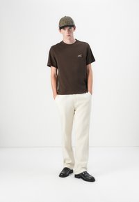 Brun kortermet t-skjorte med en liten logo, kombinert med kremfargede bukser og svarte slip-on sko. Modell med en rutete caps.