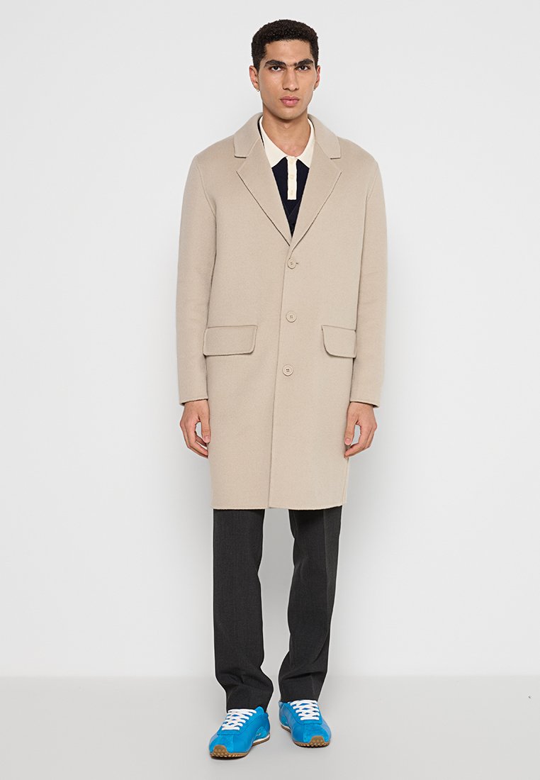 Beige Wolle-Mantel mit Reverskragen, vorderen Knöpfen und zwei Taschen, kombiniert mit dunklen Hosen und leuchtend blauen Turnschuhen.
