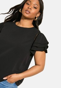 Nainen, jolla on päällään musta pusero, jossa on kerrokselliset lyhyet hihat ja kultaiset renkaakorvakorut, käsi taskussa, yksinkertaista vaaleaa taustaa vasten.