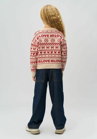 Pulover tricotat cu un model roșu și crem, prezentând inimi și textul "IUBIREA ESTE IUBIRE", asortat cu pantaloni largi din denim albastru și teniși bej.