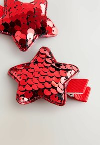 Punased tähtkujulised klambrid, kaetud läikivate litritega, koos riba silmusega kinnitamiseks ja musta tagapinnaga. Sile tekstuur koos peegeldavate aktsentidega.
