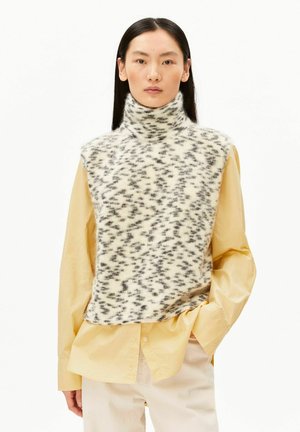 Jeune femme aux longs cheveux foncés portant un col roulé sans manches, doux, blanc moucheté de noir, par-dessus une chemise ample jaune pâle et un pantalon clair.