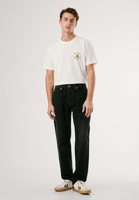 Camiseta de algodón blanca con un pequeño gráfico en el pecho, combinada con jeans de mezclilla oscura y zapatillas blancas con acentos beige.
