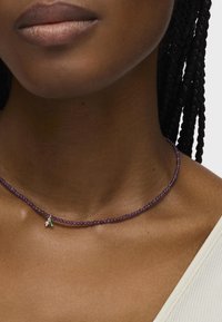 Collana di perline viola con un delicato ciondolo che presenta un piccolo accento verde. I fili sono lisci e uniformemente distanziati, poggiando delicatamente sulla pelle.