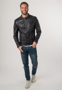 Zwarte leren jas met doorgestikte schouders, ritssluitingen en een aansluitend ontwerp. Draagts over een wit shirt en gecombineerd met blauwe jeans.