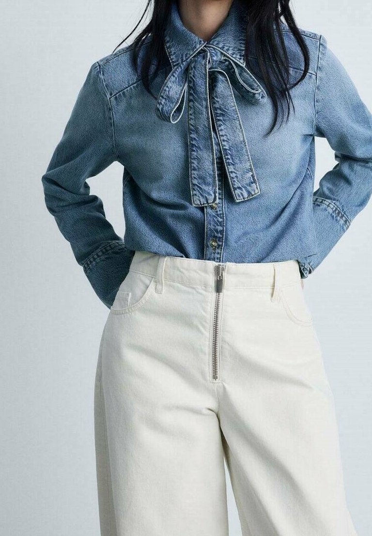 Femme portant une chemise en denim bleu avec un col noué en forme de nœud, rentrée dans un pantalon blanc taille haute à fermeture éclair frontale. Mains sur les hanches.