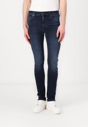 Jean skinny bleu foncé en denim, présentant un design classique à cinq poches et une légère décoloration. Porté avec des baskets blanches.