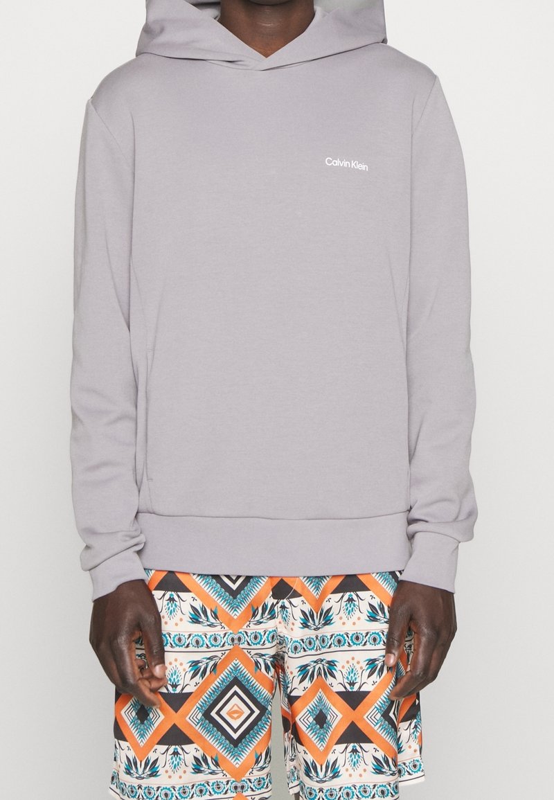 Sudadera gris claro fabricada en tejido suave, con un bolsillo frontal, puños acanalados y un sutil logo de Calvin Klein. Combinada con pantalones cortos con estampado.