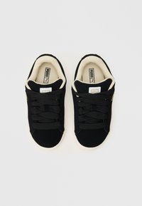 Juoda suede medžiagos sportbačiai su tekstūruotu paviršiumi, juodais raišteliais, balta padu ir auksiniais prekės ženklo akcentais šonuose.