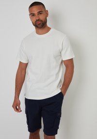 Weißes, geripptes T-Shirt mit kurzen Ärmeln, kombiniert mit marineblauen Cargo-Shorts mit Seitentaschen und strukturiertem Stoff. Das Model steht mit neutralem Gesichtsausdruck.