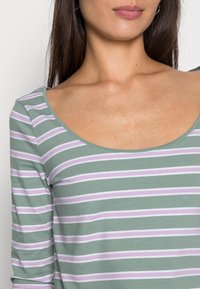 Långärmad topp i mjuk grön tyg med horisontella ränder i lavendel och vitt. Har en bred båtringning och är figurnära.