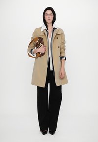 Palton bej tip trench cu un design cu două rânduri de nasturi, cămașă albă dedesubt, pantaloni negri cu croi larg, ținând o geantă maro cu model.
