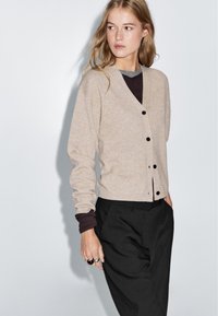 Beige ullcardigan med V-ringning, svarta knappar, långa ärmar, lager på en mörk topp, i kombination med svarta byxor. Slät textur.