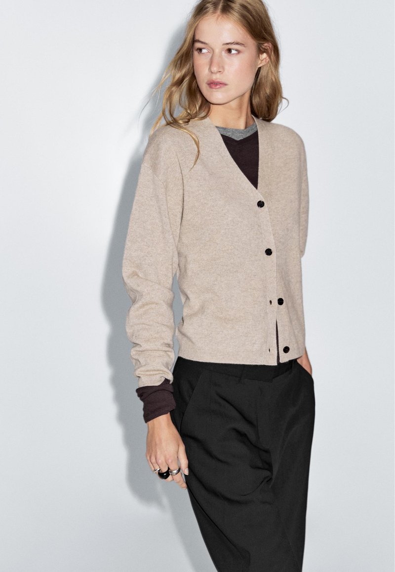 Beige ullcardigan med V-ringning, svarta knappar, långa ärmar, lager på en mörk topp, i kombination med svarta byxor. Slät textur.