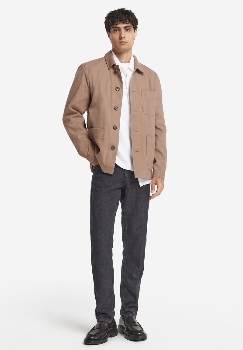 Ljusbrun canvasjacka med knapplåsningssystem och bröstficka, lagd över en vit skjorta, tillsammans med mörka denimbyxor och svarta skor.