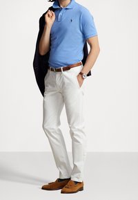 Jasnoniebieska koszulka polo, białe spodnie, brązowy skórzany pasek, brązowe zamszowe półbuty. Model trzyma granatową kurtkę, prezentuje zegarek na nadgarstku.