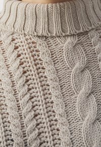 Pull en tricot beige avec un col à côtes haut, des motifs en cable détaillés et un design à mailles ouvertes, mettant en valeur des textures variées.