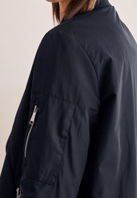 Kobieta w czarnej kurtce bombowcu z zamkniętą kieszenią na górnej części ramienia, widocznej od środkowej części pleców do okolicy ramienia.