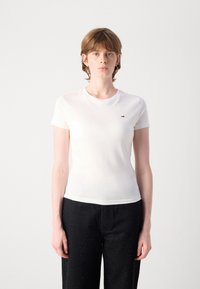 T-shirt en coton blanc avec un col rond, des manches courtes et un petit logo sur la poitrine gauche, associé à un pantalon noir avec des détails subtils.