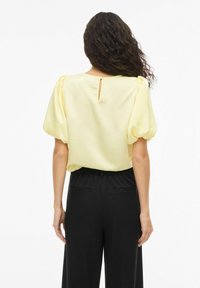 Blouse jaune à manches bouffantes, texture lisse, détail dos avec ouverture en forme de goutte. Associée à un pantalon noir taille haute avec poches latérales.