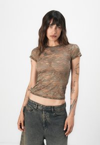Camiseta ajustada de tela transparente con estampado de camuflaje, con mangas cortas y cuello redondo, combinada con pantalones cortos de mezclilla oscura.