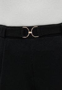 Μαύρο παντελόνι από ύφασμα με μεταλλική χρυσή διπλή αγκράφα ζώνης και υφασμένη μέση, τοποθετημένο σε φωτεινό ριμπ φόντο.