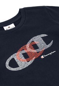 Camiseta de algodón azul marino con logotipos interconectados en blanco y rojo con un diseño texturizado; mangas cortas y cuello redondo. Etiqueta de Champion visible.