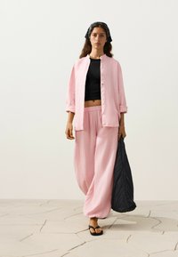 Femme portant un pantalon rose ample et une chemise rose assortie boutonnée par-dessus un crop top noir, tenant un grand sac noir et portant des sandales noires.