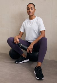 Juodi sportiniai bateliai, balta marškinėliai ir violetinės leggins. Vandens butelis rankose. Sėdi ant juodų mankštos kamuolių prieš šviesią foną.
