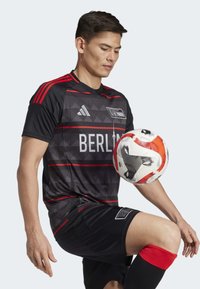 Maillot de football noir avec des accents rouges et des motifs géométriques, affichant le texte "BERLIN". Le joueur équilibre un ballon de football sur son genou, portant des chaussettes rouges et noires.