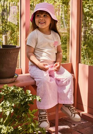 Fiatal lány rózsaszín sapkában és nadrágban, aki egy rózsaszín padon ül egy üvegházban, mosolyogva oldalra néz.