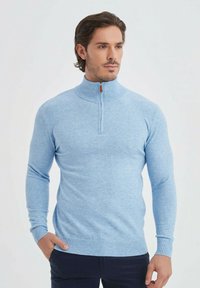 Pull en tricot bleu clair avec un col montant et une fermeture éclair sur un quart. Poignets et ourlet côtelés, texture lisse, coupe décontractée.
