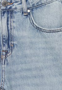 Gros plan sur un jean en denim bleu clair montrant la poche avant, la passants de ceinture, le bouton métallique et les détails de surpiqûres orange.