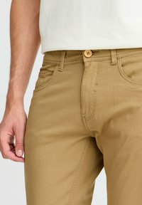 Khaaki chinos valmistettu puuvillasekoitteesta, suoran leikkauksen, etutaskujen ja napitushelman kanssa, jossa on teksturoitu pinta.