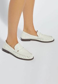 White leather loafers with a pleated design and rounded toe. Features a low, brown rubber sole and a decorative strap.