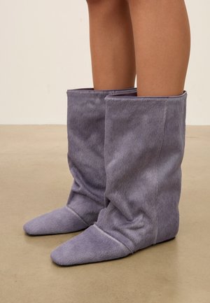 Personne portant des bottes violettes souples, amples, mi-mollet avec un bout arrondi, debout sur un sol beige lisse.