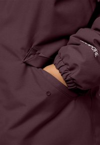 Giacca bordeaux con polsini elasticizzati, una tasca laterale e tessuto morbido con finitura opaca. Include un dettaglio del logo discreto.