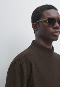 Bruine geribbelde trui met opstaande kraag en een gestructureerde afwerking. Het onderwerp draagt donkere zonnebril en is vastgelegd vanuit een licht zijaanzicht.