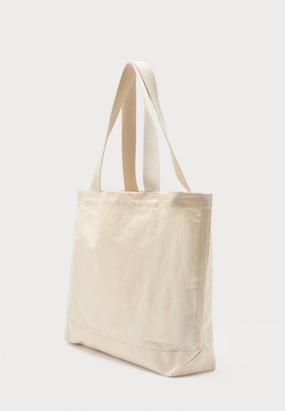 Sac cabas en toile beige avec double sangle d'épaule, base rectangulaire et design simple et dépouillé sur fond blanc.