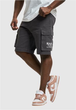 Osoba nosící tmavě šedé cargo šortky s bočními kapsami a bílo-růžové tenisky Nike, stojící před jednobarevným pozadím.