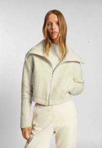 Kraftig krämfärgad jacka med struktur, stor krage och dragkedja. Långa ribbade ärmar kompletterar vida krämfärgade byxor.