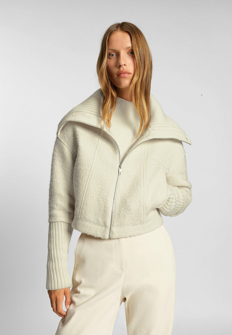 Kraftig krämfärgad jacka med struktur, stor krage och dragkedja. Långa ribbade ärmar kompletterar vida krämfärgade byxor.