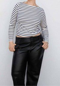 Personne portant une chemise blanche à manches longues avec des rayures horizontales noires et un pantalon en cuir noir taille haute, les mains dans les poches.