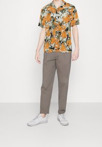 Mand iført kortærmet skjorte med orangemønstret, brune slim-fit bukser og hvide sneakers, der står foran en hvid baggrund.